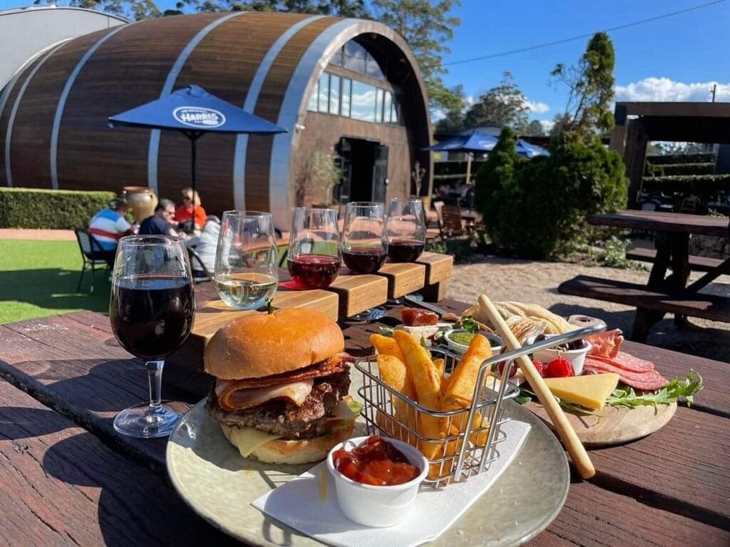 Outdoor patio scene with a cheeseburger, fries, and a small dipping sauce on a wooden table, plus several wine glasses and a side board of snacks in the background.