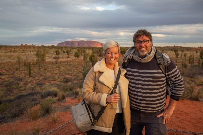 Smiling couple in desert at sunset, with red soil, sparse vegetation, and Uluru in the background.