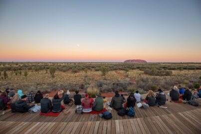 Group of people seated on wooden benches outdoors at dusk, facing a desert landscape with a distant rock formation (Uluru) under a colorful sunset sky.