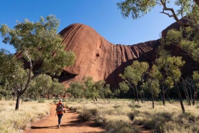 Hiker in an orange jacket walks a red dirt trail toward a tall ribbed red rock formation with trees and a blue sky overhead.