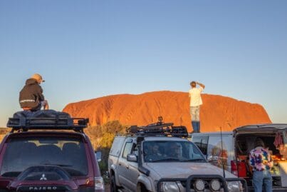 Two people stand on top of parked SUVs with bike racks, gazing at a large red rock formation against a clear blue sky.
