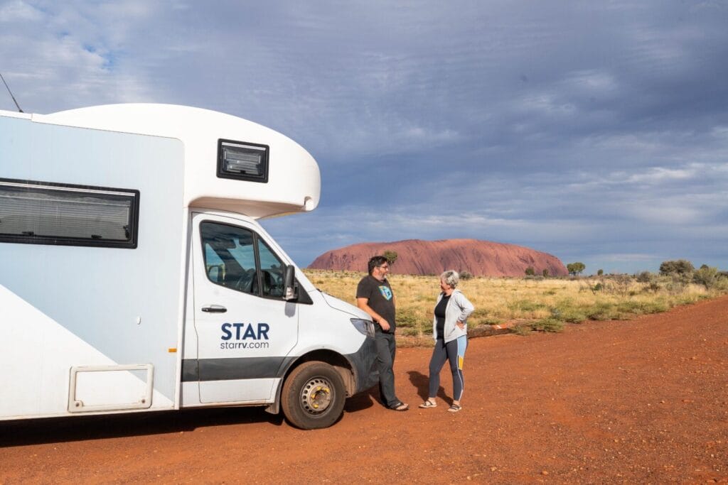 uluru - Londoner In Sydney White motorhome with STAR branding parked on red dirt desert, two people talking by the cab, Uluru rock in the background under a cloudy sky.