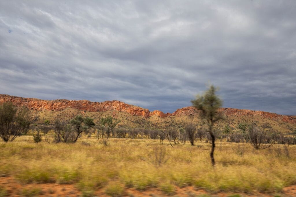 west-macdonnell-ranges-road - Londoner In Sydney Desert landscape with red rock hills under a cloudy sky and scattered trees and brush in the foreground.